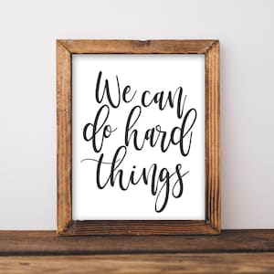 May include: A framed print with a white background and black calligraphy that reads "We can do hard things". The frame is made of brown wood.
