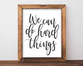 May include: A framed print with a white background and black calligraphy text that reads "We can do hard things". The frame is made of brown wood.