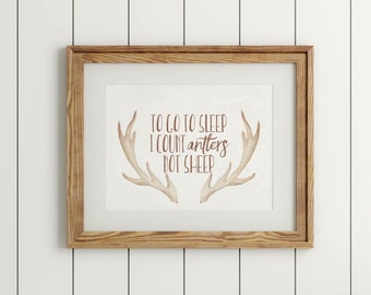 May include: A framed print with a brown wooden frame and a white background. The print features a pair of antlers with the text "To go to sleep I count antlers not sheep" in brown lettering.