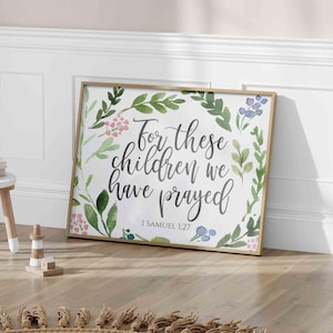Può includere: Una stampa incorniciata con un design a corona di fiori e il testo "For these children we have prayed" con il riferimento biblico "I Samuel 1:27". La stampa è appesa a un muro sopra un tappeto marrone con frange.