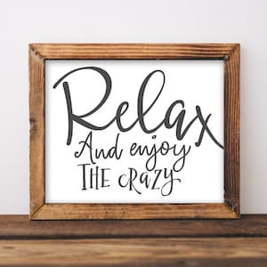 May include: A framed sign with the text "Relax and enjoy the crazy" in a black cursive font on a white background. The frame is made of brown wood.