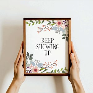 May include: A framed print with a floral design and the text "KEEP SHOWING UP" in black lettering. The frame is brown wood.