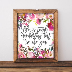 May include: A framed print with a watercolor floral border in shades of pink, purple, and green. The text "Life is tough my darling but so are you" is written in elegant black script on a white background. The frame is made of brown wood.