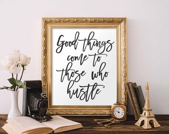 May include: A gold framed print with the quote "Good things come to those who hustle" in black calligraphy on a white background. The print is sitting on a wooden table with a book, a camera, a clock, and a miniature Eiffel Tower.