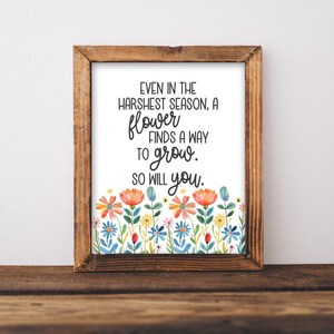 May include: A framed print with a wooden frame. The print features the text "Even in the harshest season, a flower finds a way to grow. So will you." A colorful floral border of watercolor flowers decorates the bottom of the print.
