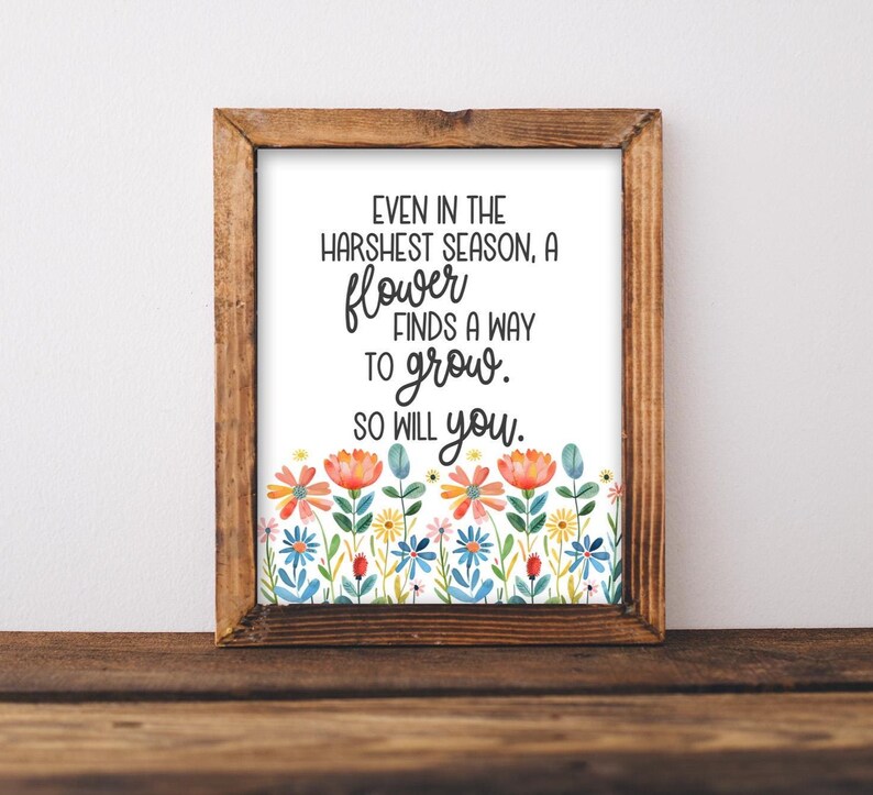 May include: A framed print with a wooden frame. The print features the text "Even in the harshest season, a flower finds a way to grow. So will you." A colorful floral border of watercolor flowers decorates the bottom of the print.
