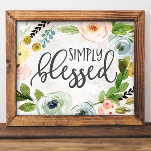 May include: A wooden framed sign with a white background and watercolor floral design. The sign says "SIMPLY blessed" in black script.