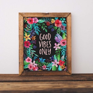 Puede incluir: Una impresión enmarcada con fondo negro y una corona floral colorida que rodea el texto "GOOD VIBES ONLY" en letras rosas.