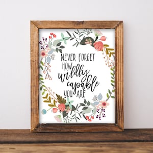 Op de afbeelding: Een ingelijste print met een bloemenkransontwerp. De print bevat de tekst "NEVER FORGET HOW wildly capable YOU ARE" in zwart schrift. De krans bevat roze, blauwe en groene bloemen en bladeren. Het frame is gemaakt van bruin hout.