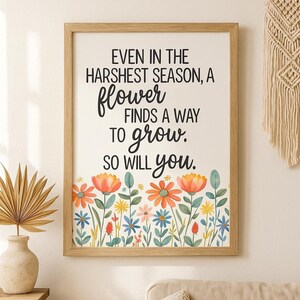 May include: A framed print with the quote "EVEN IN THE HARSHEST SEASON, A flower FINDS A WAY TO grow. SO WILL you." The print features a floral border with orange, blue, and yellow flowers. The frame is made of light-colored wood.