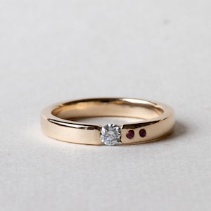 May include: A gold ring featuring a central clear gemstone and two small red gemstones. The ring has a minimalist design. The ring is set against a white background.