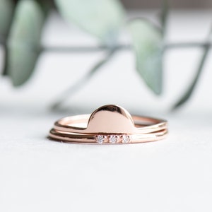 Puede incluir: Un conjunto de dos anillos de oro rosa. El anillo superior presenta una forma de semicírculo, mientras que el anillo inferior tiene una fila de tres pequeños diamantes.