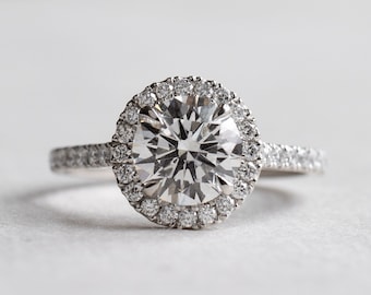 Könnte beinhalten: Ein silberner Verlobungsring mit einem großen, runden, klaren Diamanten in der Mitte, der von einem Heiligenschein aus kleineren Diamanten umgeben ist. Die Ringschiene ist ebenfalls mit kleineren Diamanten besetzt.