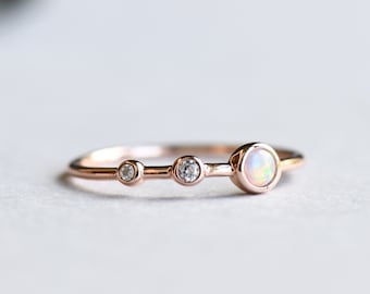 May include: A delicate rose gold ring with three round gemstones. The center stone is a larger, white opal, flanked by two smaller, clear diamonds.