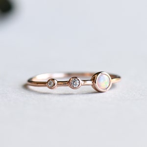 Puede incluir: Un delicado anillo de oro rosa con tres piedras redondas. La piedra central es una opala blanca más grande, flanqueada por dos diamantes claros más pequeños.