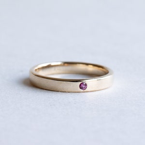 May include: A simple gold band ring with a single small pink gemstone set in the center.