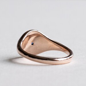 May include: A rose gold signet ring with a small blue gemstone set in the center of the top. The ring is smooth and has a rounded shape.