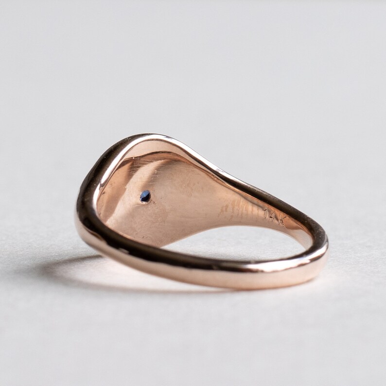 May include: A rose gold signet ring with a small blue gemstone set in the center of the top. The ring is smooth and has a rounded shape.