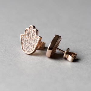 May include: A pair of rose gold earrings featuring a hamsa hand design. The earrings are set with small, clear crystals.