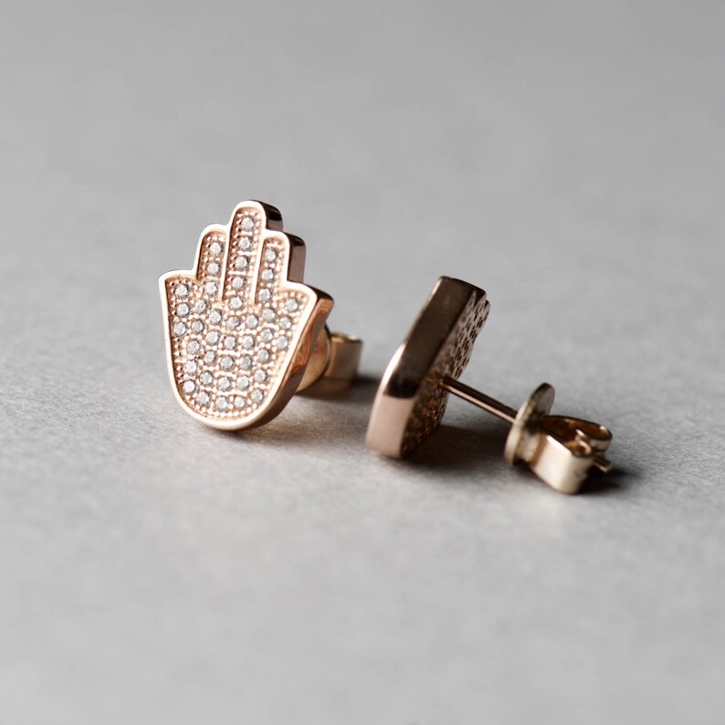 May include: A pair of rose gold earrings featuring a hamsa hand design. The earrings are set with small, clear crystals.