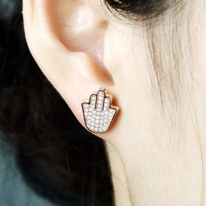 May include: Rose gold earrings featuring a hamsa hand design, fully encrusted with clear cubic zirconia stones.