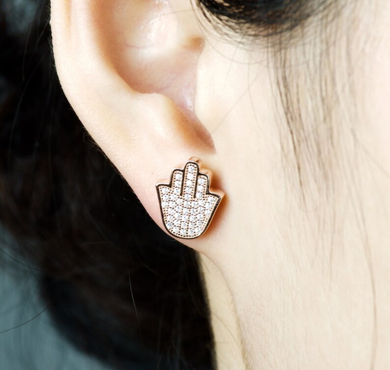 May include: Rose gold earrings featuring a hamsa hand design, fully encrusted with clear cubic zirconia stones.