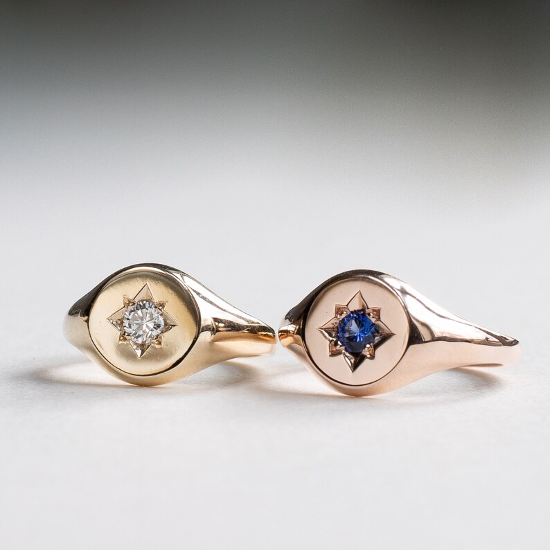May include: Two gold signet rings with a star design. One ring has a clear stone in the center of the star, and the other ring has a blue stone in the center of the star.
