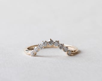 May include: A curved gold ring set with multiple small, clear gemstones. The ring's design features a gentle arch of sparkling stones, creating a delicate and elegant appearance. The ring is shown against a soft, neutral background.