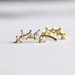 May include: A pair of gold ear cuffs with a curved design and clear cubic zirconia stones.