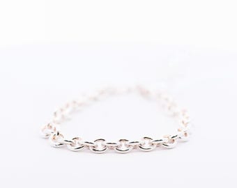 Puede incluir: Una pulsera de cadena plateada con una serie de eslabones redondeados interconectados. La pulsera se muestra sobre un fondo blanco, resaltando su superficie pulida y reflectante. La cadena es de diseño sencillo y elegante.
