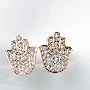 May include: A pair of gold-tone earrings featuring a hamsa hand design. Each earring is set with clear cubic zirconia stones.