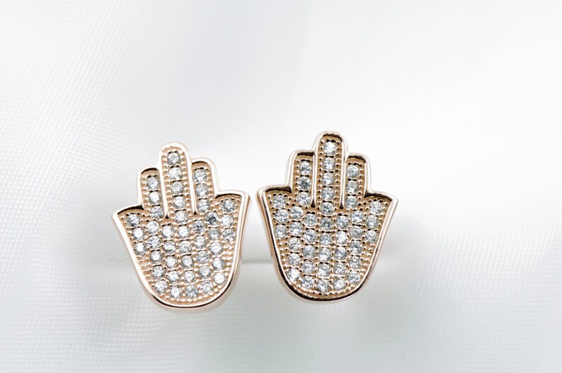 May include: A pair of gold-tone earrings featuring a hamsa hand design. Each earring is set with clear cubic zirconia stones.