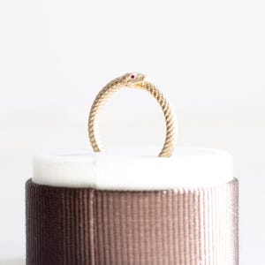 May include: A gold-colored ring in the shape of a snake, with a red gemstone eye. The snake's body is textured, and the ring is displayed on a white surface, set against a neutral background.