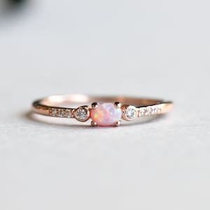 Könnte beinhalten: Ein zarter Ring aus Roségold mit einem rosafarbenen Opal-Edelstein und kleinen klaren Steinen, die in einer Bandfassung sitzen.