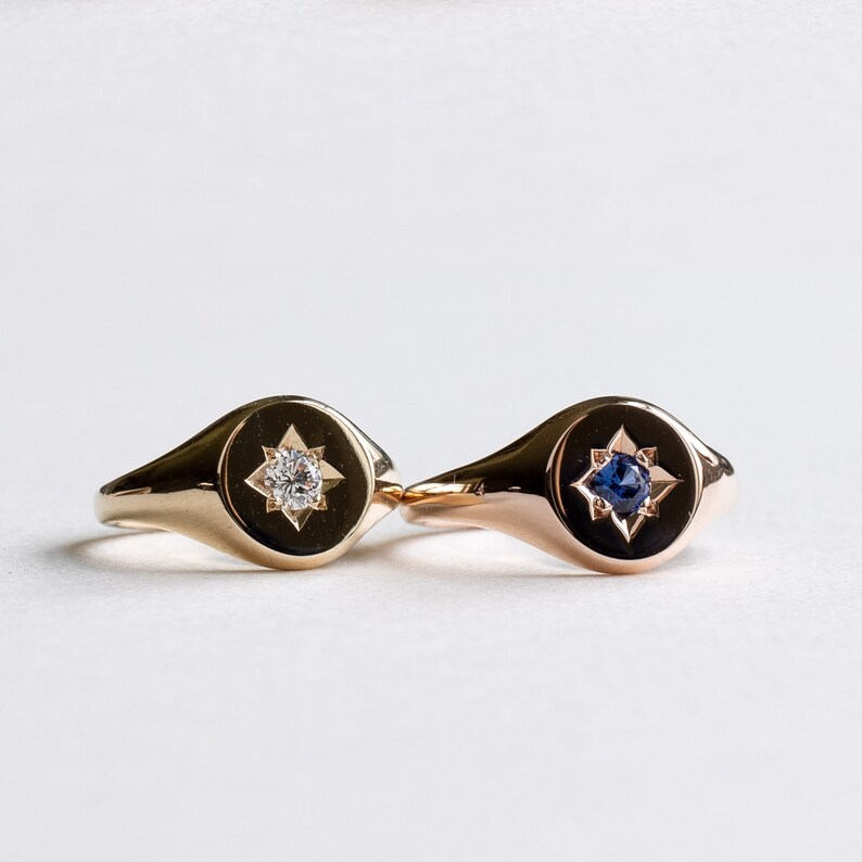 May include: Two gold signet rings with a star design. One ring has a clear stone in the center of the star, and the other ring has a blue stone in the center of the star.