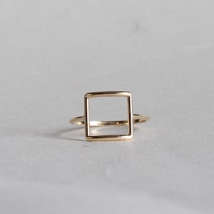 Może przedstawiać: Minimalistyczny złoty pierścionek z kwadratowym wzorem. Pierścionek ma cienką obrączkę i pusty kwadrat pośrodku. Pierścionek wykonany jest z błyszczącego, złotego metalu i jest umieszczony na neutralnym tle.