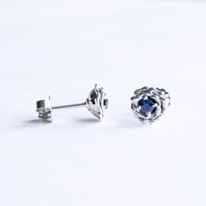 May include: A pair of silver stud earrings with a blue gemstone in the center of each earring. The earrings are designed to look like a flower.