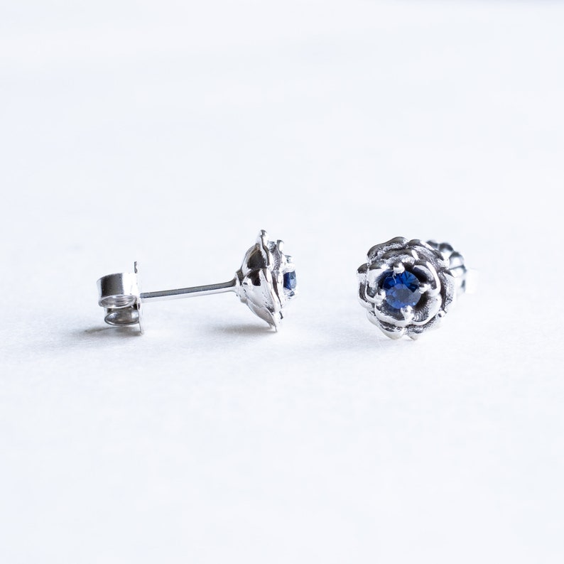 May include: A pair of silver stud earrings with a blue gemstone in the center of each earring. The earrings are designed to look like a flower.