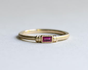 May include: A delicate gold band ring with a small, rectangular, red ruby gemstone set in the center.