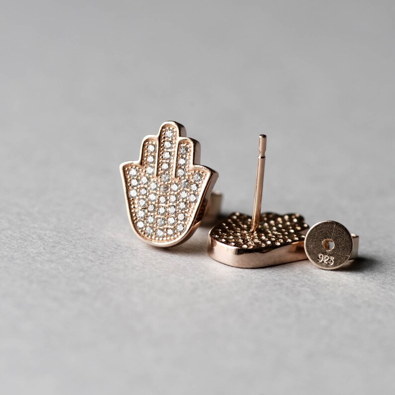 May include: A pair of rose gold earrings featuring a hamsa hand design. The earrings are set with small, clear cubic zirconia stones.