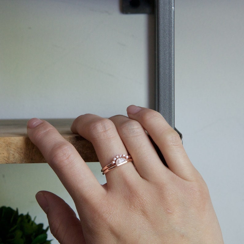 May include: A close-up of a hand wearing two rose gold rings. The top ring features a half-moon shaped diamond with smaller diamonds around the edge. The bottom ring is a simple band.