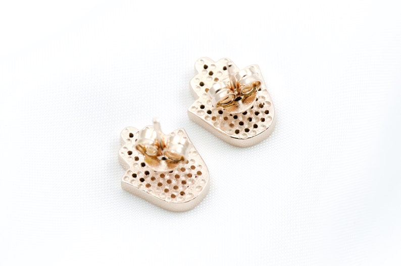 May include: A pair of gold-toned earrings featuring a hamsa hand design. The earrings are made of metal and have a post back fastening.