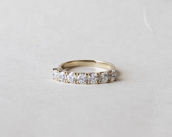 Può includere: Un anello in oro con sette pietre preziose rotonde e trasparenti. L'anello è appoggiato su una superficie bianca e la fascia ha una finitura lucida. Le pietre sono distanziate uniformemente e riflettono la luce.