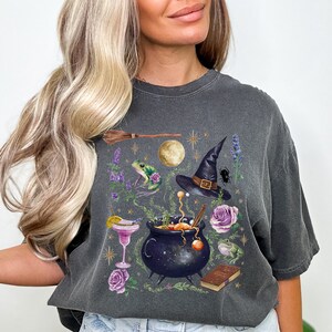 May include: Dark grey t-shirt featuring a whimsical witch-themed design. The graphic includes a cauldron, witch hat, frog, broom, moon, flowers, and a cocktail. The design is in shades of purple, green, and gold.