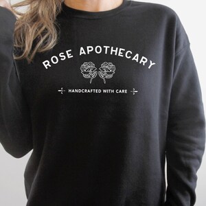 May include: Black crewneck sweatshirt with the text "ROSE APOTHECARY" in white, arched above two white rose illustrations. Below, the text "HANDCRAFTED WITH CARE" is centered, with a decorative line on each side.