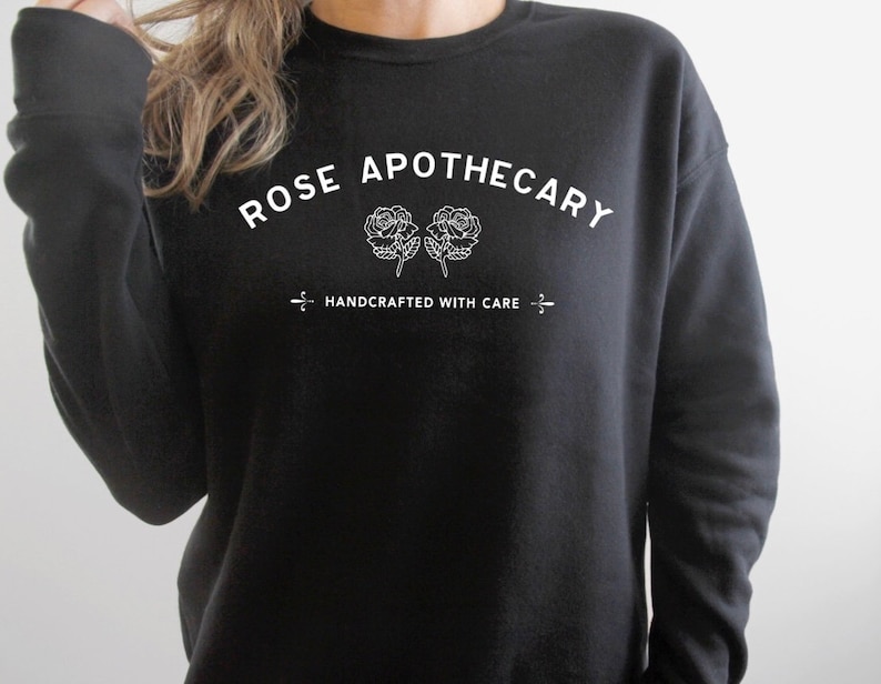 May include: Black crewneck sweatshirt with the text "ROSE APOTHECARY" in white, arched above two white rose illustrations. Below, the text "HANDCRAFTED WITH CARE" is centered, with a decorative line on each side.