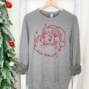 May include: A heather gray sweatshirt with a red Santa Claus face graphic. The long-sleeved top is hanging on a wooden hanger. The Santa Claus graphic includes a beard, hat, and stars. The sleeves are cuffed.