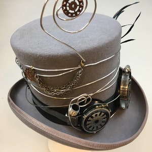 May include: A gray steampunk top hat with silver wire accents, a small copper gear, and attached goggles. The hat features a black band and decorative chain, with a feather detail.