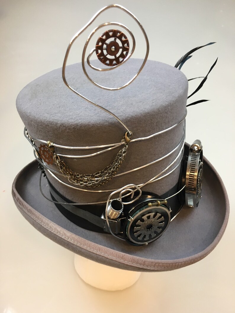 May include: A gray steampunk top hat with silver wire accents, a small copper gear, and attached goggles. The hat features a black band and decorative chain, with a feather detail.