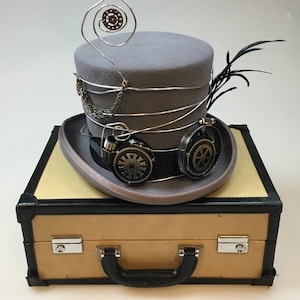 May include: A gray steampunk top hat with silver wire accents, a chain, and goggles. The hat features a black band and is adorned with a feather. It rests on a tan and black suitcase with a handle and silver clasps.
